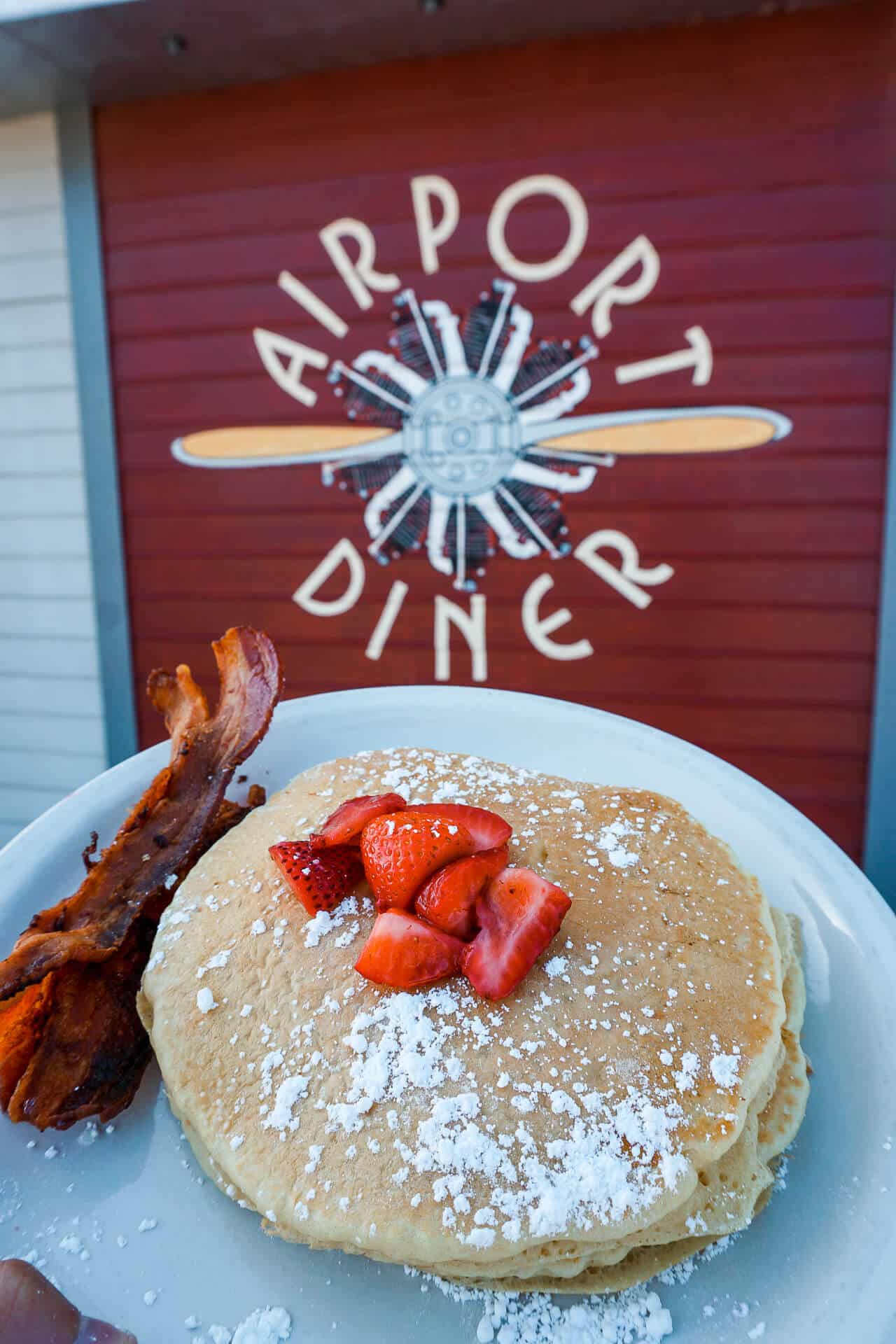 A stack of pancakes with sliced strawberries on top and a side of bacon with the Airport Diner logo in the background (from Fredericksburg, TX)