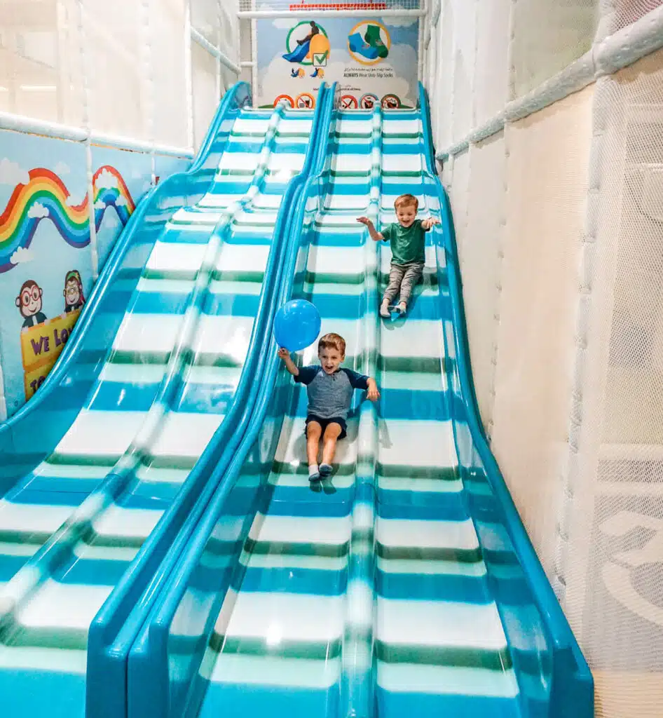 Two toddler boys racing down the blue slides at Cheeky Monkey, an indoor playground in Dallas.