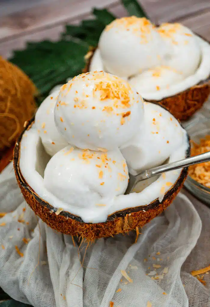 A spoon going into a coconut shell filled with three scoops of homemade Coconut Sorbet with toasted coconut on top.