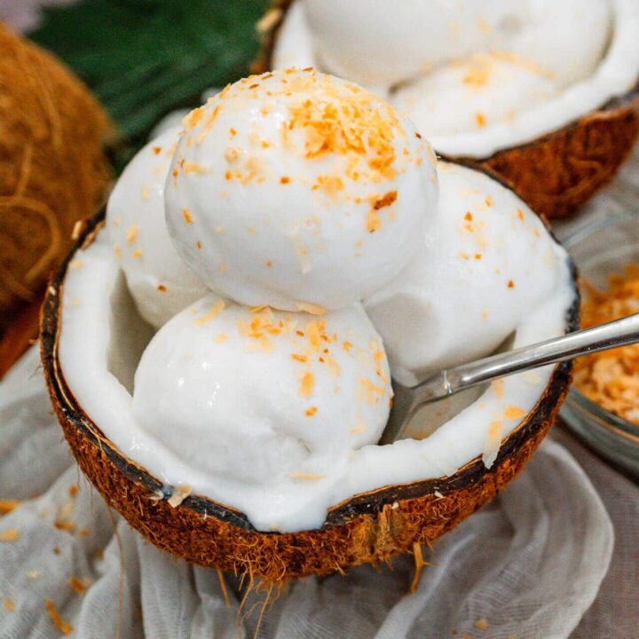 A spoon going into a coconut shell filled with three scoops of homemade Coconut Sorbet with toasted coconut on top.