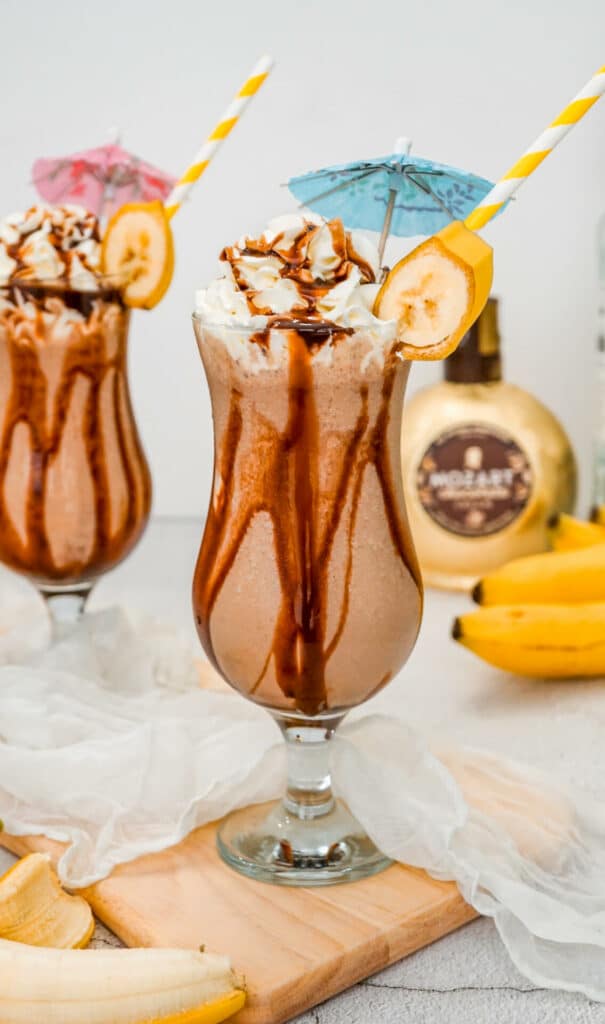 A hurricane glass filled with a chocolate-banana Dirty Monkey Frozen Cocktail with chocolate drizzle on the sides and topped with whipped cream. A bottle of Mozart chocolate liqueur in the back.