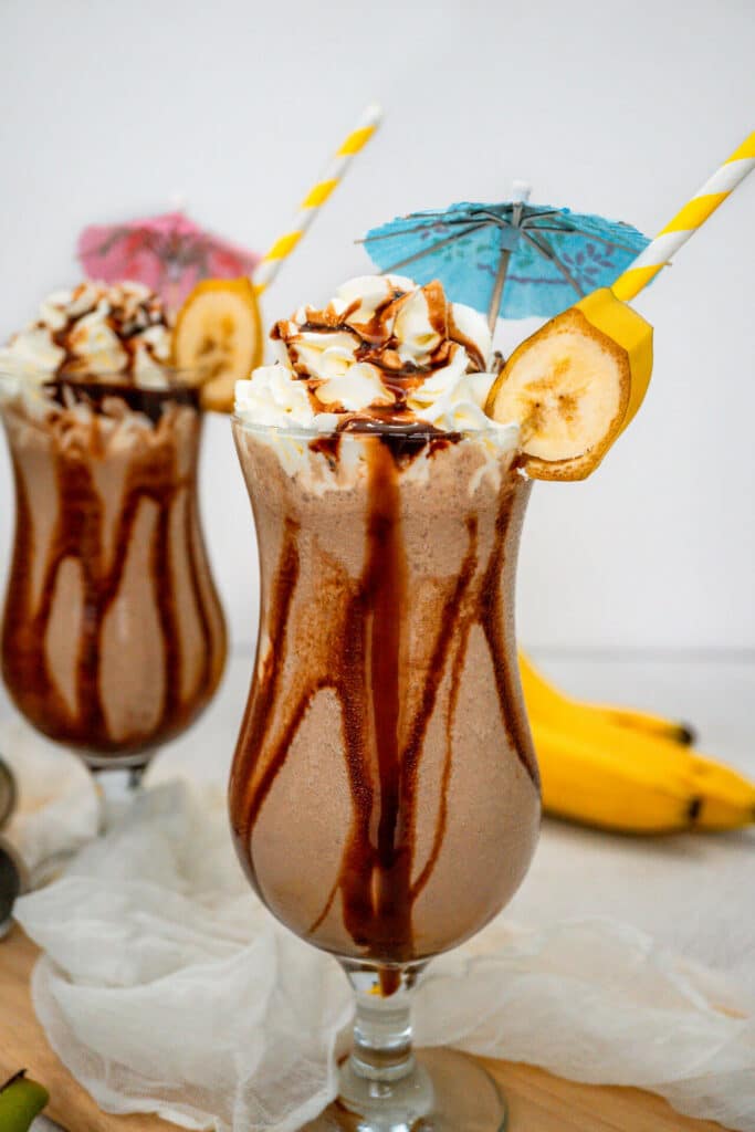 Two hurricane glasses filled with a Dirty Monkey Frozen Cocktail with chocolate drizzle on the sides and topped with whipped cream.