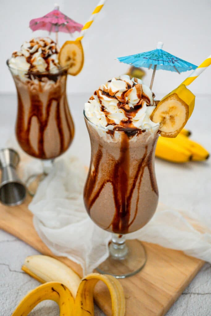 Two hurricane glasses on a wooden board filled with a Dirty Monkey Frozen Cocktail with chocolate drizzle on the sides, topped with whipped cream, umbrellas, a yellow paper straw and a slice of banana.