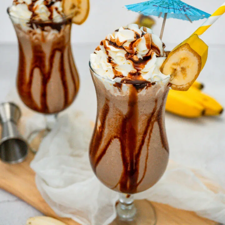 Two hurricane glasses on a wooden board filled with a Dirty Monkey Frozen Cocktail with chocolate drizzle on the sides, topped with whipped cream, umbrellas, a yellow paper straw and a slice of banana.