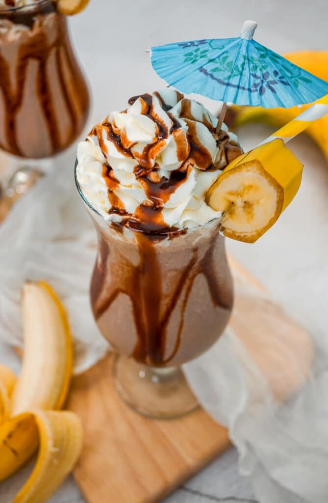 A hurricane glass filled with a Dirty Monkey Frozen Cocktail with chocolate drizzle on the sides, topped with whipped cream, a blue umbrella and a slice of banana.