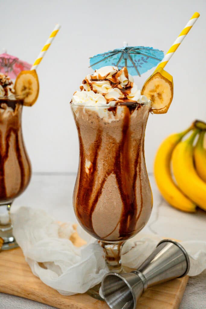 A hurricane glass on a wooden board filled with a Dirty Monkey Frozen Cocktail with chocolate drizzle on the sides and topped with whipped cream—and a jigger laying sideways next to the drink.