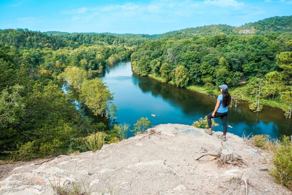 A woman standing on a bluff looking out into the Mountain Fork River in Broken Bow, Oklahoma. The trail is Cedar Bluff and hiking it is one of the best things to do in Broken Bow.