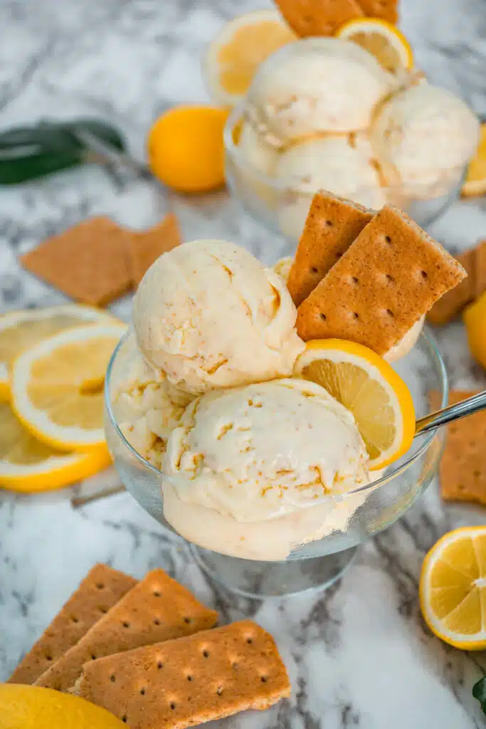 Scoops of Lemon Pie Ice Cream in a clear glass bowl with graham crackers and a slice of lemon on top.