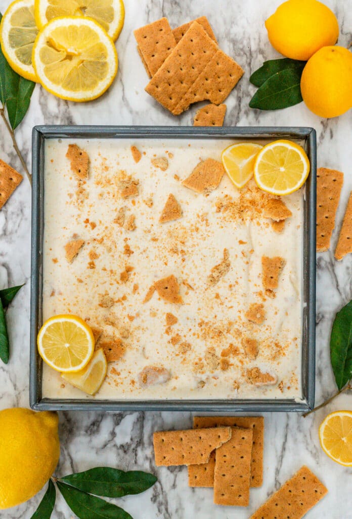 A square dish filled with Lemon Pie Ice Cream with shards of graham crackers on top and slices of lemon on the corners.