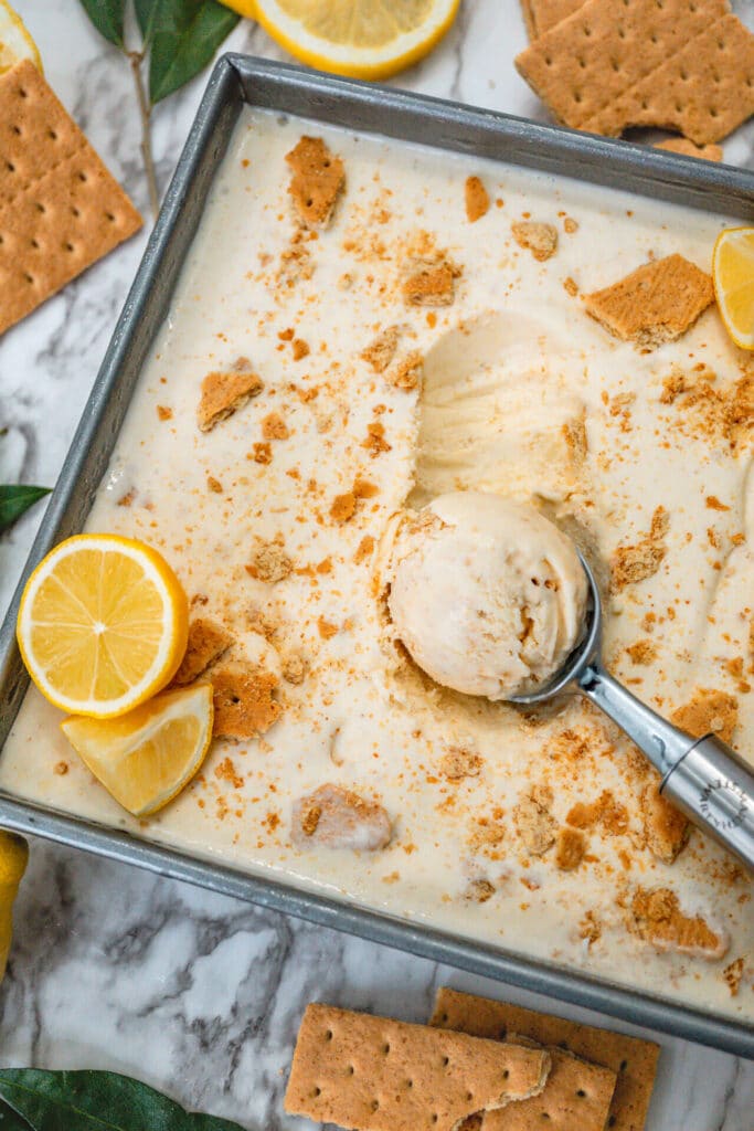 An ice cream scoop going into a square pan of homemade Lemon Pie Ice Cream with shards of graham cracker and sliced lemon on top.