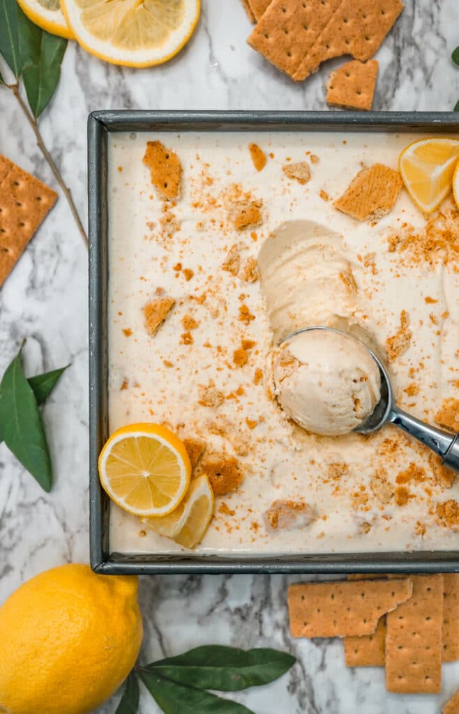 A birdseye view of a Lemon Pie Ice Cream in a square dish with graham cracker shards and sliced lemon on top. There is also an ice scream scoop scooping a ball of Lemon Pie Ice Cream. 