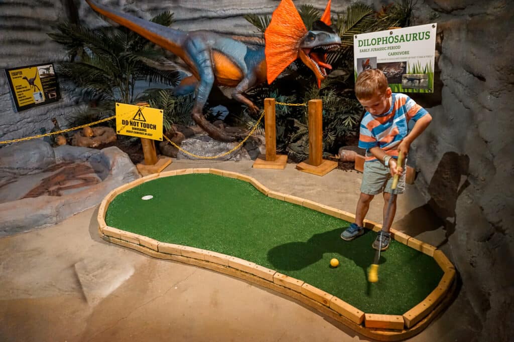 A young boy hitting a golf ball on a small putting green with a dinosaur in the background (location: Beavers Bend Mining Company in Broken Bow, Oklahoma).