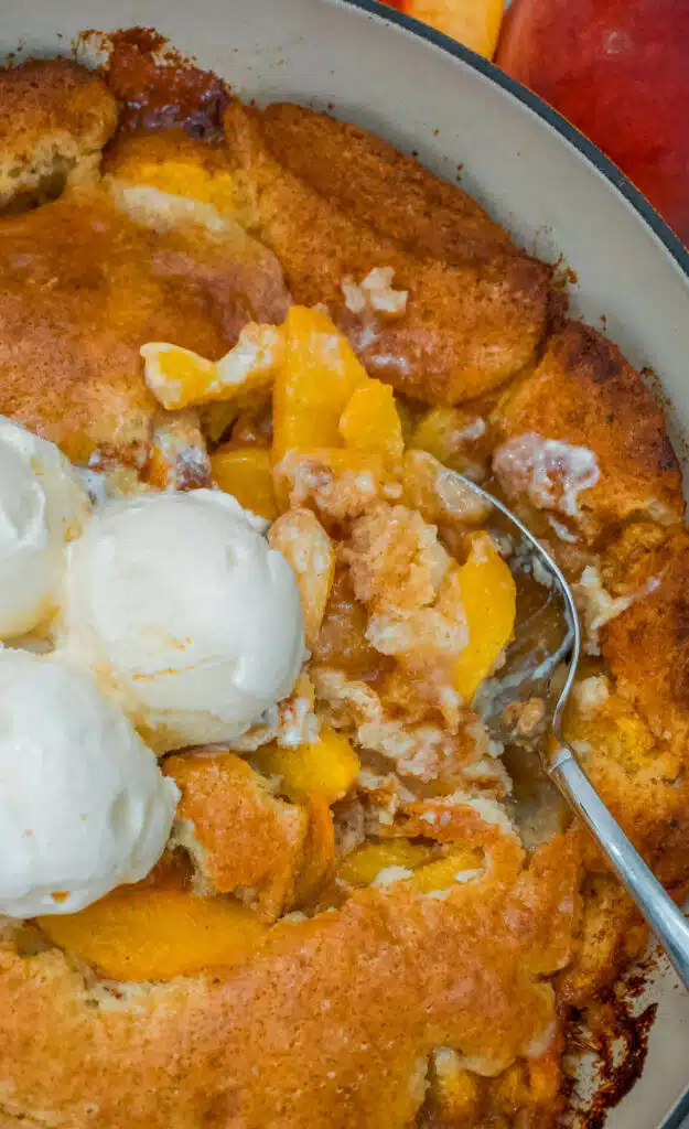 A spoon digging into a juicy Southern-style Dutch oven peach cobbler with scoops of vanilla ice cream on top.