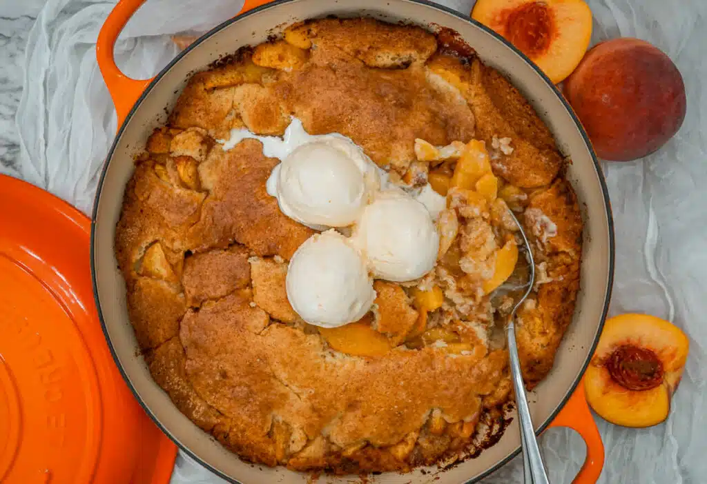 A Southern-style peach cobbler baked in an orange Dutch oven. A spoon is digging into the cobbler with three scoops of vanilla ice cream on top. 