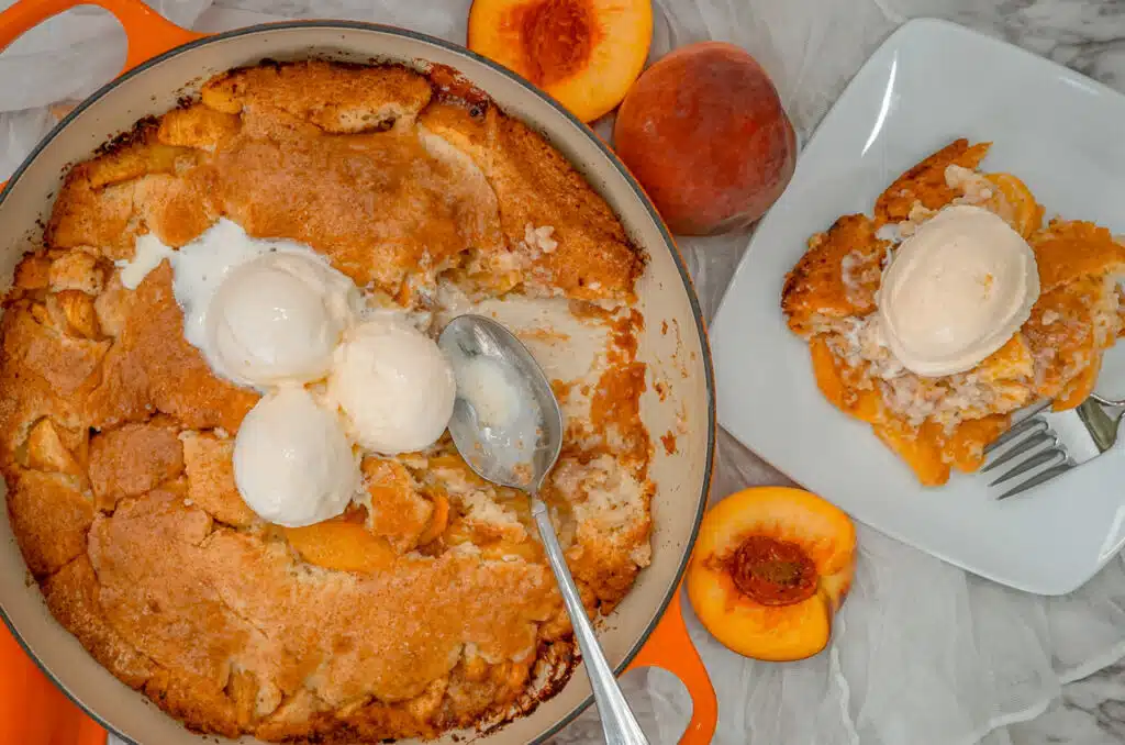 A Southern-style peach cobbler baked in a Dutch oven with a portion missing, a spoon, and three scoops of vanilla ice cream on top. Next to it is a portion of peach cobbler on a white plate with a scoop of ice cream on top.