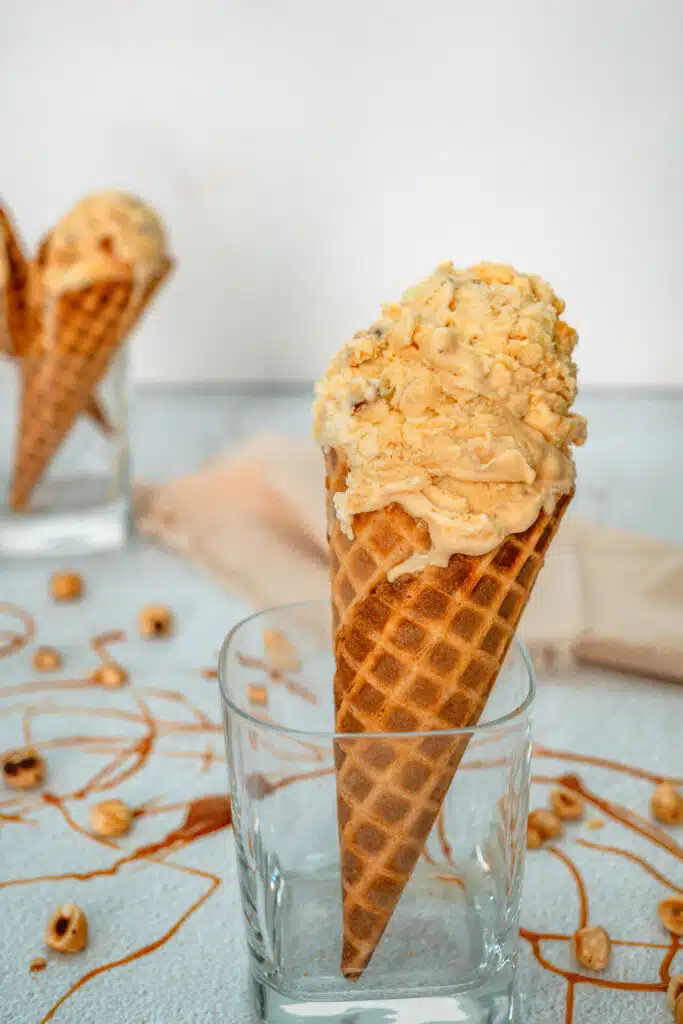 A glass holding a waffle cone filled with scoops of homemade Hazelnuts Salted Caramel Ice Cream. There is also caramel drizzle and toasted hazelnuts surrounding the glass. 