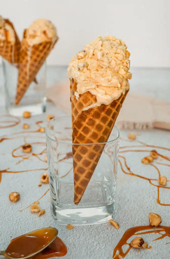 A glass holding a waffle cone filled with scoops of homemade Hazelnuts Salted Caramel Ice Cream. There is also a spoon of caramel, caramel drizzle, and toasted hazelnuts surrounding the glass. 