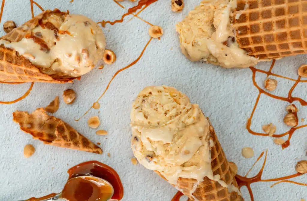 Waffle cones filled with scoops of homemade Hazelnuts Salted Caramel Ice Cream laying sporadically on a table. There is also a spoon of caramel, caramel drizzle, and toasted hazelnuts surrounding the area. 