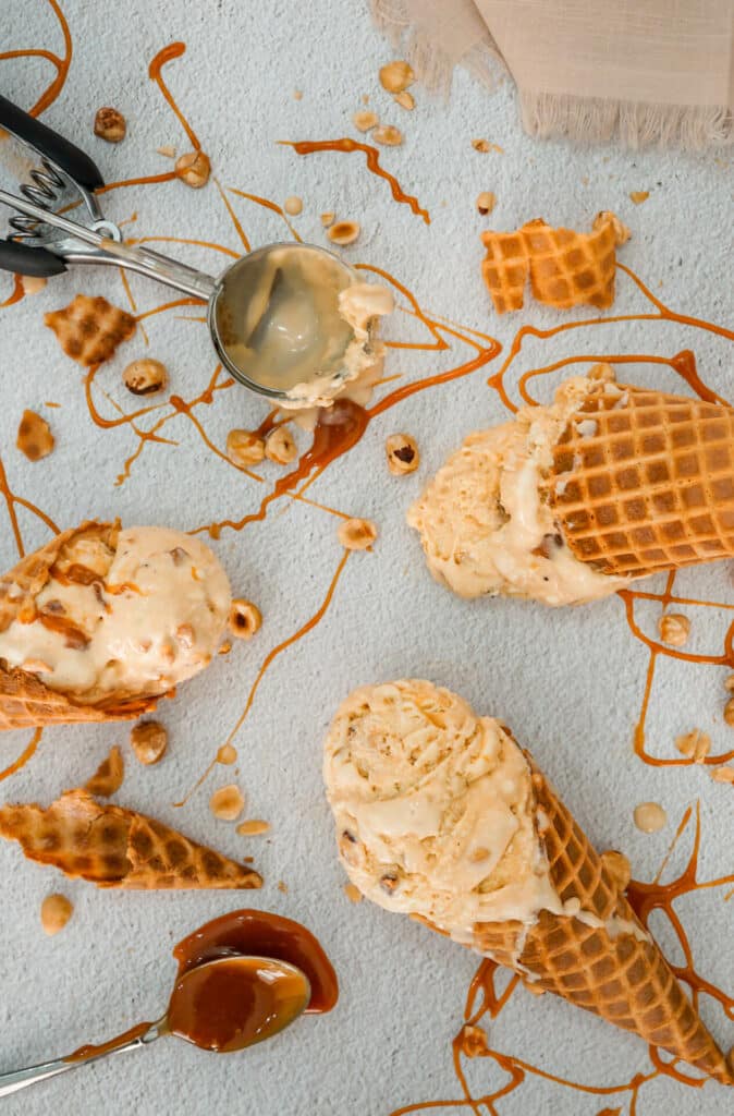 Waffle cones filled with scoops of homemade Hazelnuts Salted Caramel Ice Cream laying sporadically on a table. There is also an ice cream scoop, spoon of caramel, caramel drizzle, and toasted hazelnuts surrounding the area. 