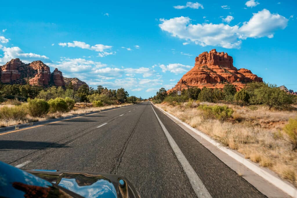 A car driving the Red Rock Scenic Byway with the iconic Bell Rock butte in view. 