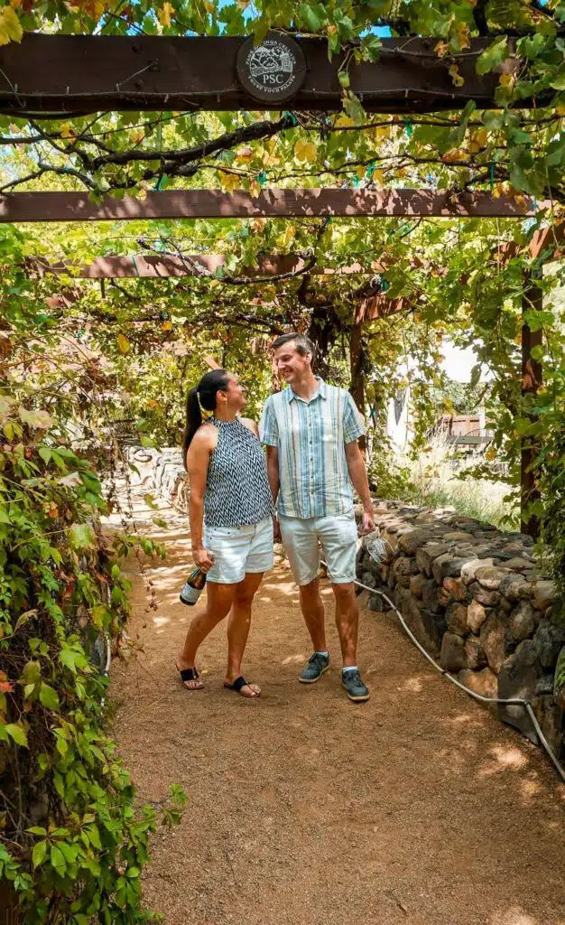 A sweet couple holding a bottle of wine and glasses walking under a vines at Page Springs Cellars. 